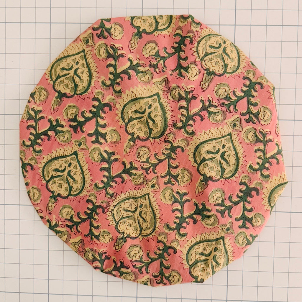 Decorative Bath hat with green and gold pattern on a pink background, placed on a grid surface.