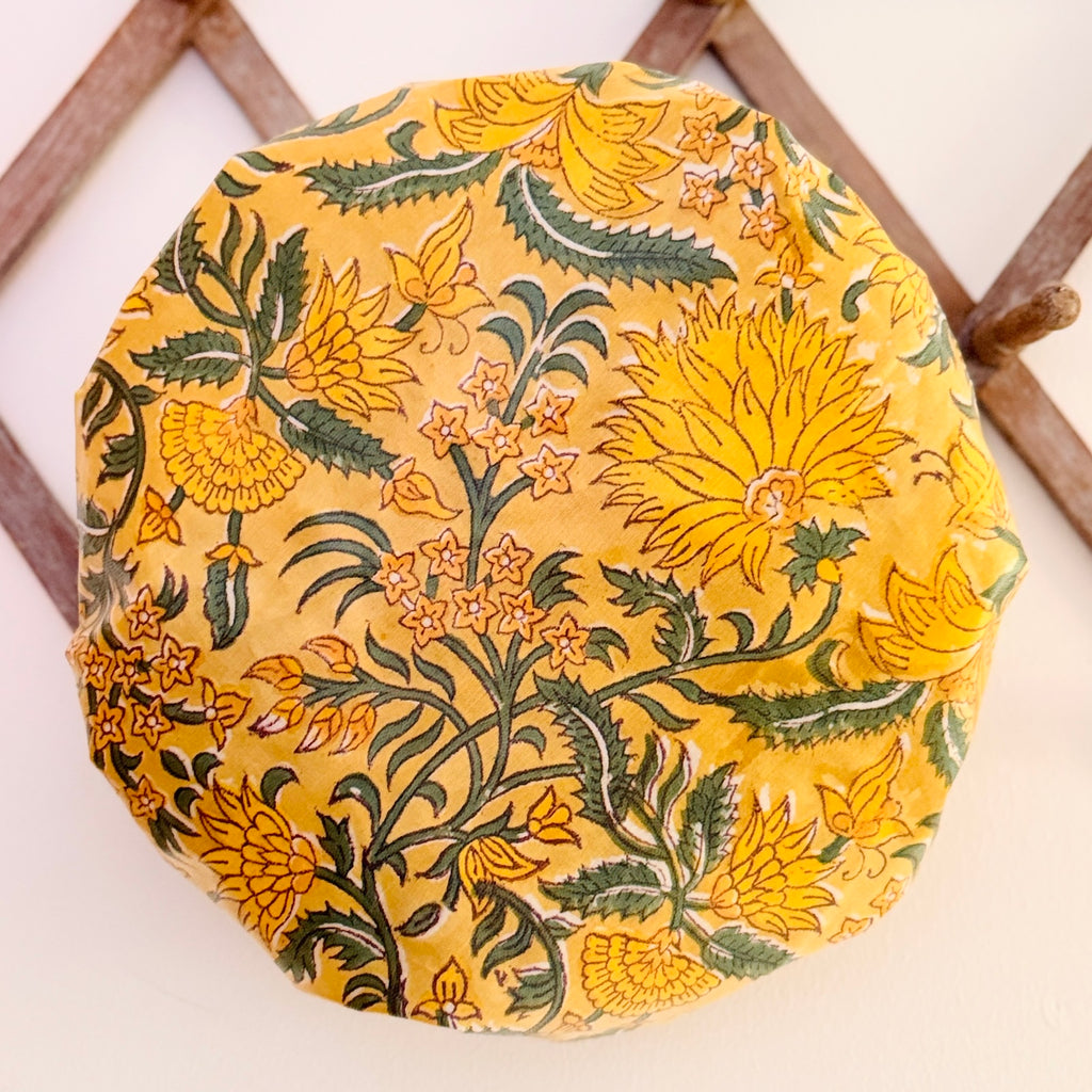 Shower Cap in vibrant yellow floral pattern with green leaves, displayed on a light-colored surface with a wooden lattice background.