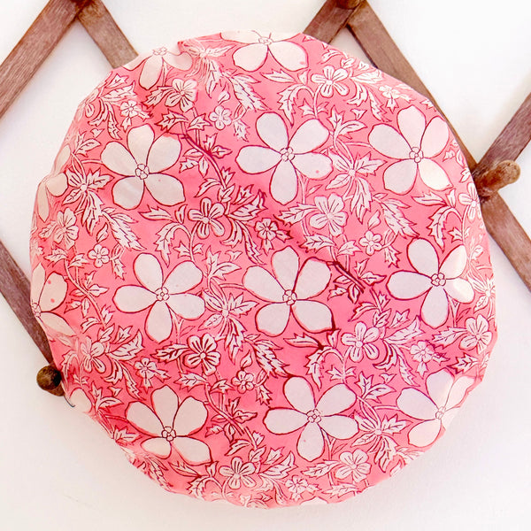 A round pink fabric shower cap with a white floral pattern, displayed on a wooden lattice against a white background.