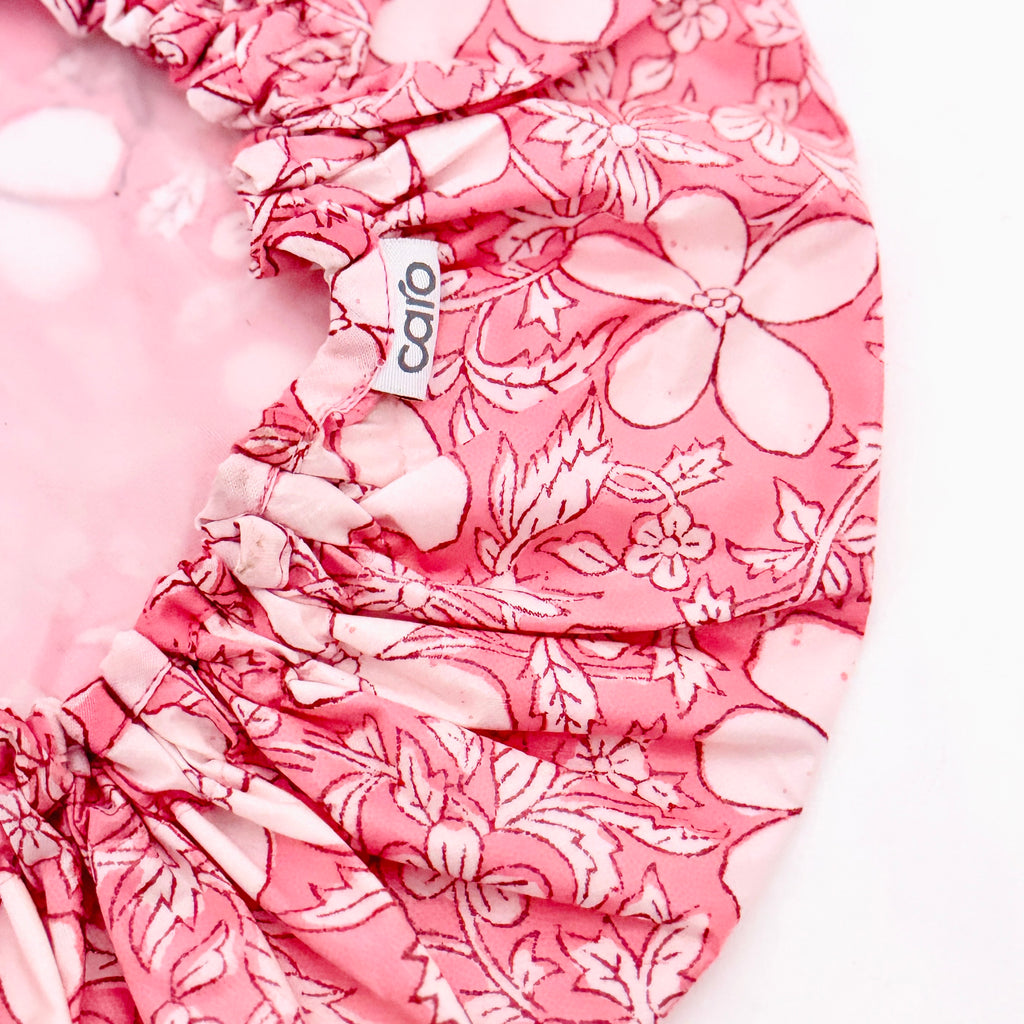 A close-up of a pink floral fabric product with gathered ruffles and a small white tag reading 'caro' on a white background.