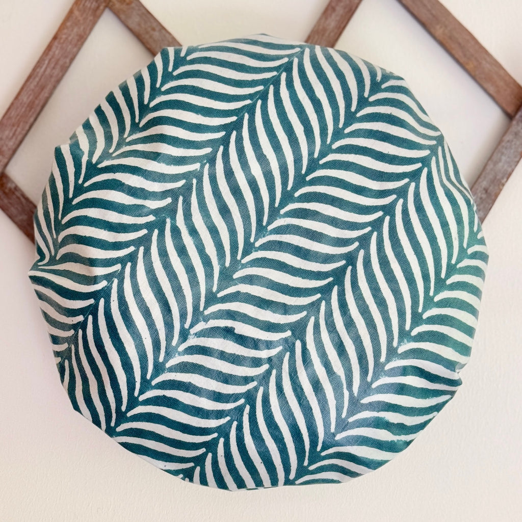 A round fabric shower cap featuring a teal and white hand-drawn leaf pattern, displayed on a light beige wall in front of a wooden geometric frame.