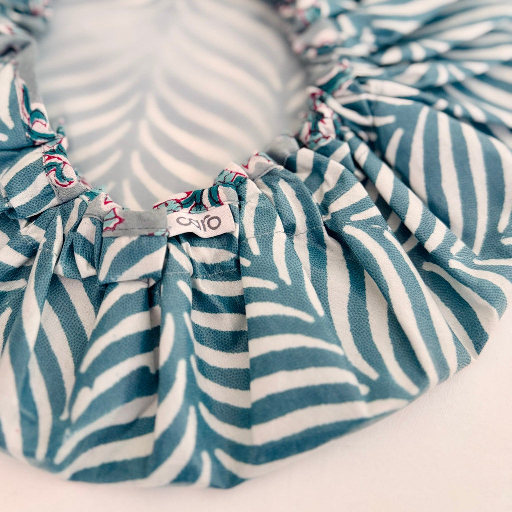 A close-up of a ruffled shower cap with a teal and white zebra-like stripe pattern, featuring a small fabric tag with the brand name 'caro'.