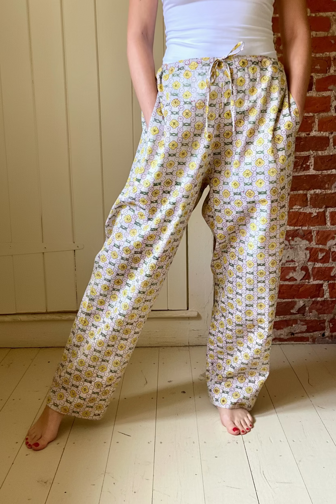 A person wearing wide legged pajama bottoms with pockets in daisy chain pattern in yellow, green, and cream colors.