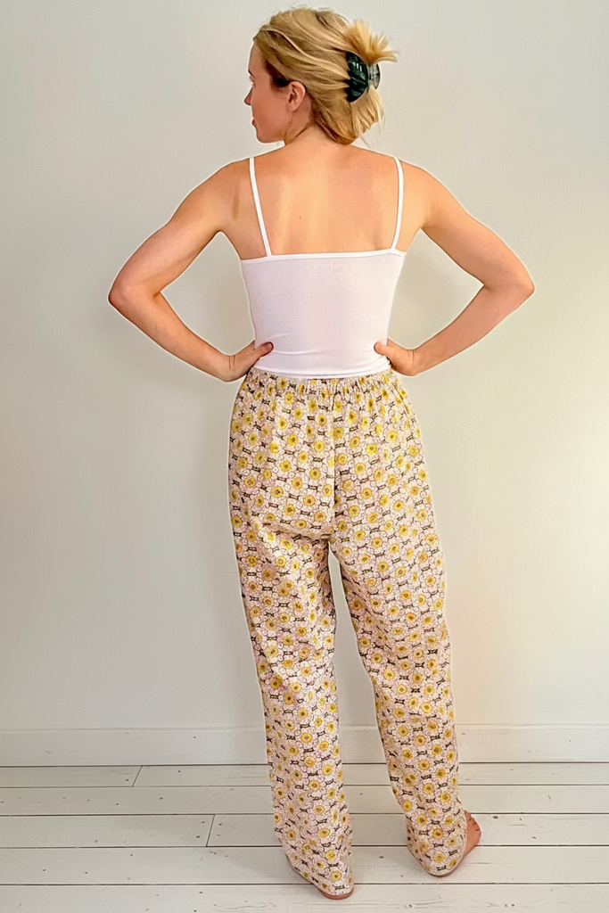 Back view of woman wearing wide legged pajama bottoms with pockets in daisy chain pattern in yellow, green, and cream colors.
