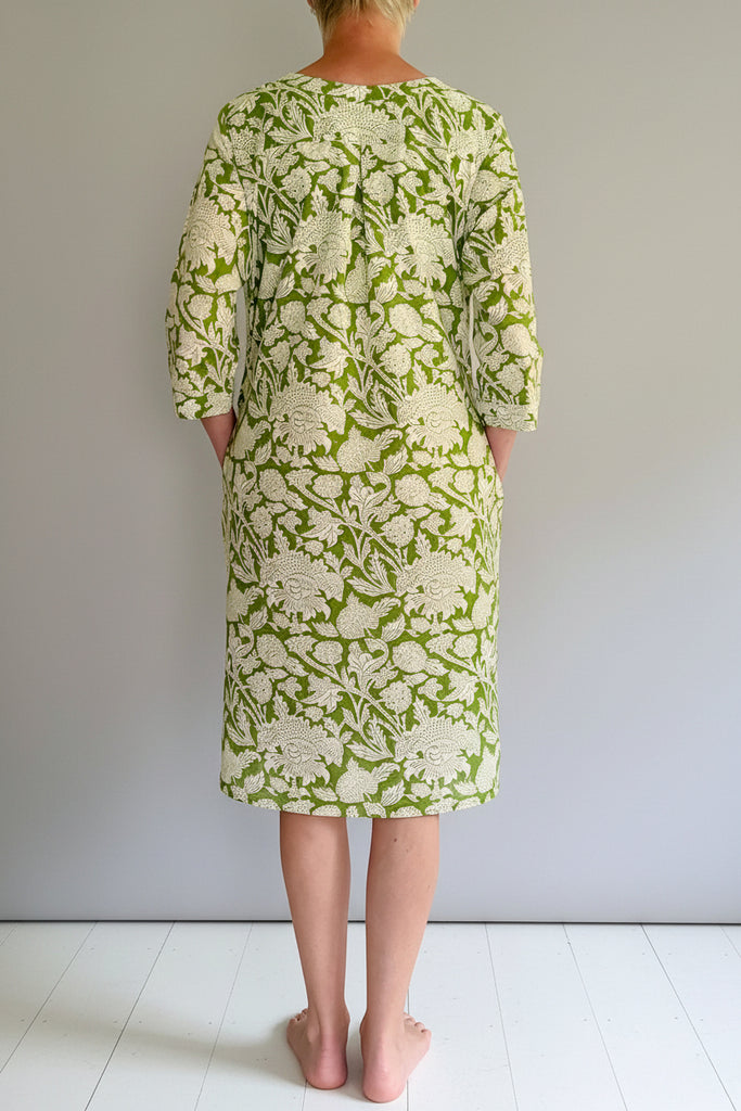Person wearing a green floral dress standing on a wooden platform against a white wall.