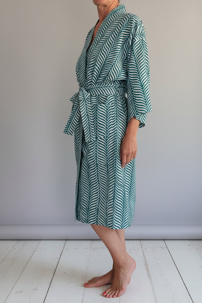 A person wearing a long-sleeve tie-front robe with a green and white wavy herringbone pattern, standing barefoot on a light wooden stool against a plain off-white wall.