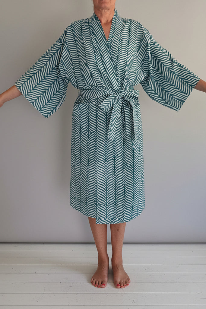 A person wearing a long-sleeved tie-front robe with a green and white chevron pattern, standing barefoot on a light-colored wooden platform against a plain white wall.