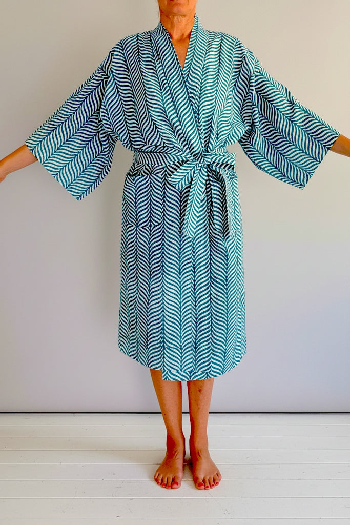 A person wearing a teal and white patterned kimono-style robe with wide sleeves, standing barefoot against a plain light gray wall and white floor.