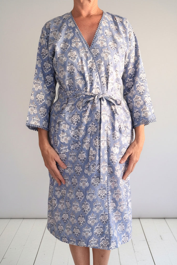 A woman wearing a light blue and white floral print wrap robe with three-quarter sleeves and a tie waist, standing against a light gray wall and white wooden floor.
