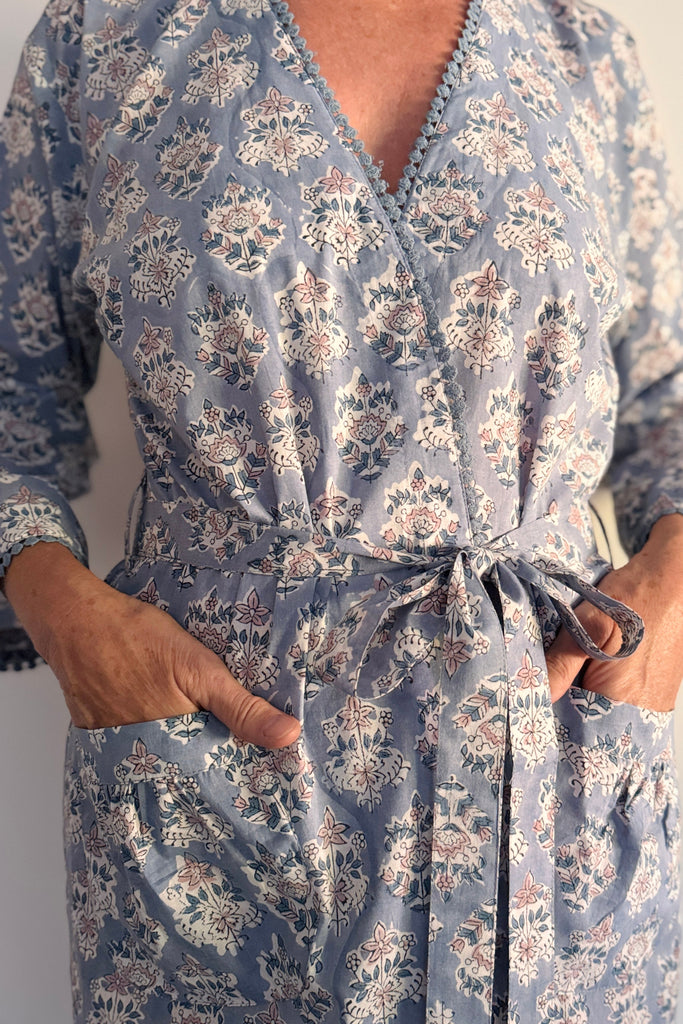 A close-up of a person wearing a dusty blue wrap-style dress with a white and beige floral block print, featuring a tie waist and pockets.