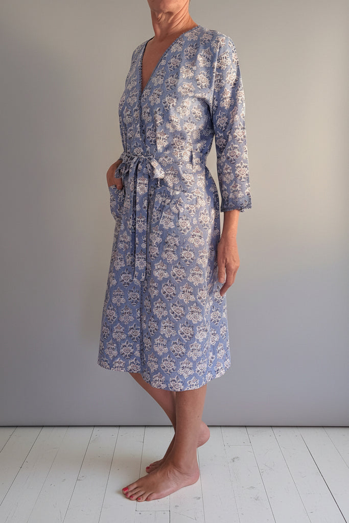 A woman models a knee-length wrap robe with a blue and white floral pattern, standing barefoot against a plain wall.