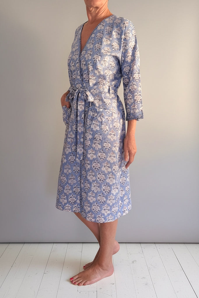 A woman modeling a knee-length wrap dress with three-quarter sleeves and a waist tie, featuring a blue and white floral pattern, standing against a neutral gray wall and white wooden floor.