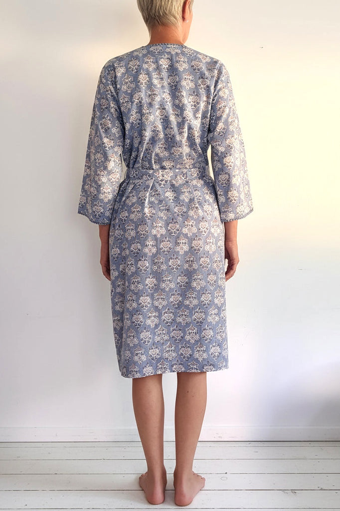 A person viewed from behind wearing a knee-length patterned robe with three-quarter sleeves and a tie waist, standing against a plain light wall.