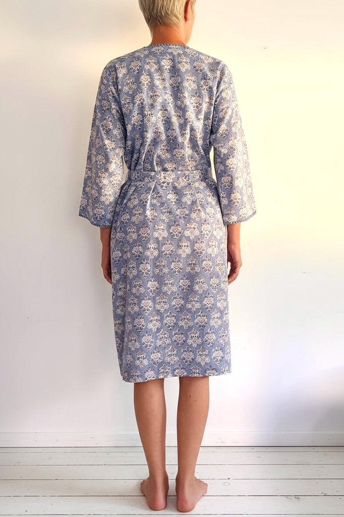A person viewed from behind wearing a knee-length patterned robe with three-quarter sleeves and a self-tie belt, standing barefoot against a plain white wall.