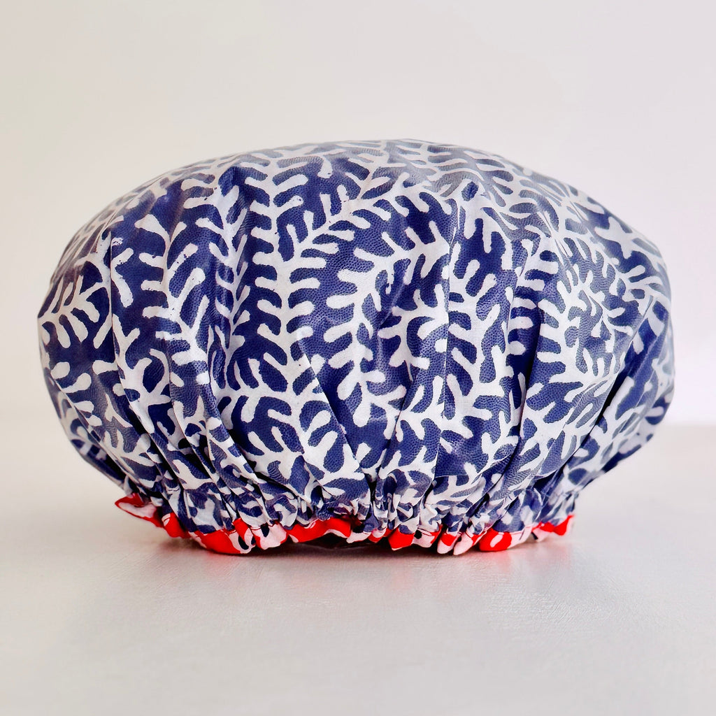 Waterproofed shower cap with a blue and white coral leaf pattern and a contrast orange ruffled trim, photographed on a neutral light beige background.