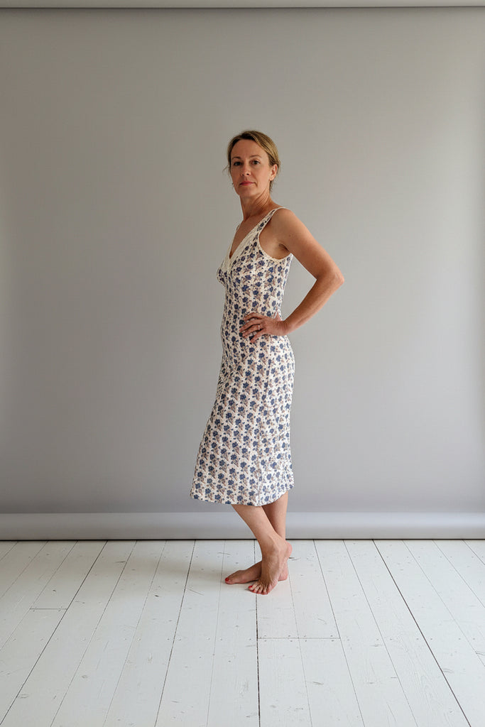 Woman wearing a floral dress standing on a wooden platform against a white wall.