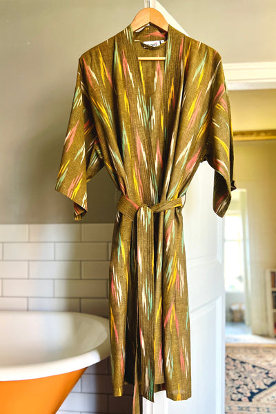 An olive Ikat woven kimono robe with multicolor patterns, displayed on a hanger in a room setting, featuring 3/4 length sleeves and a tie belt.