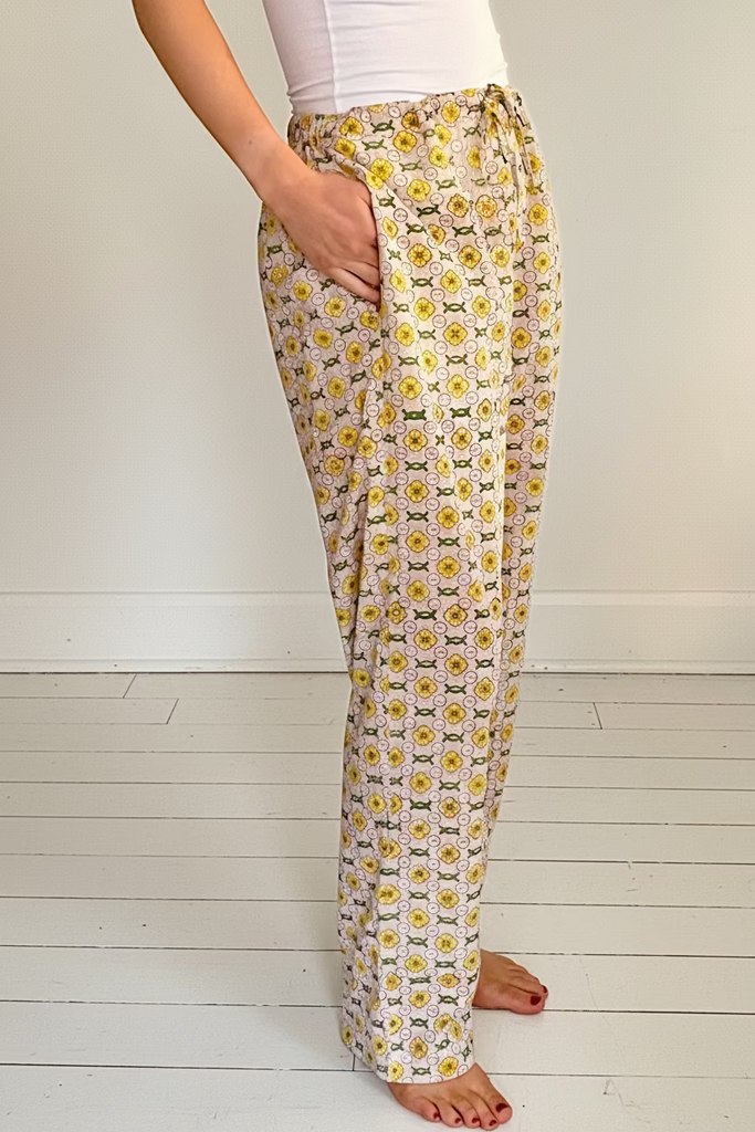 Side view of woman wearing wide legged pajama bottoms with pockets in daisy chain pattern in yellow, green, and cream colors.